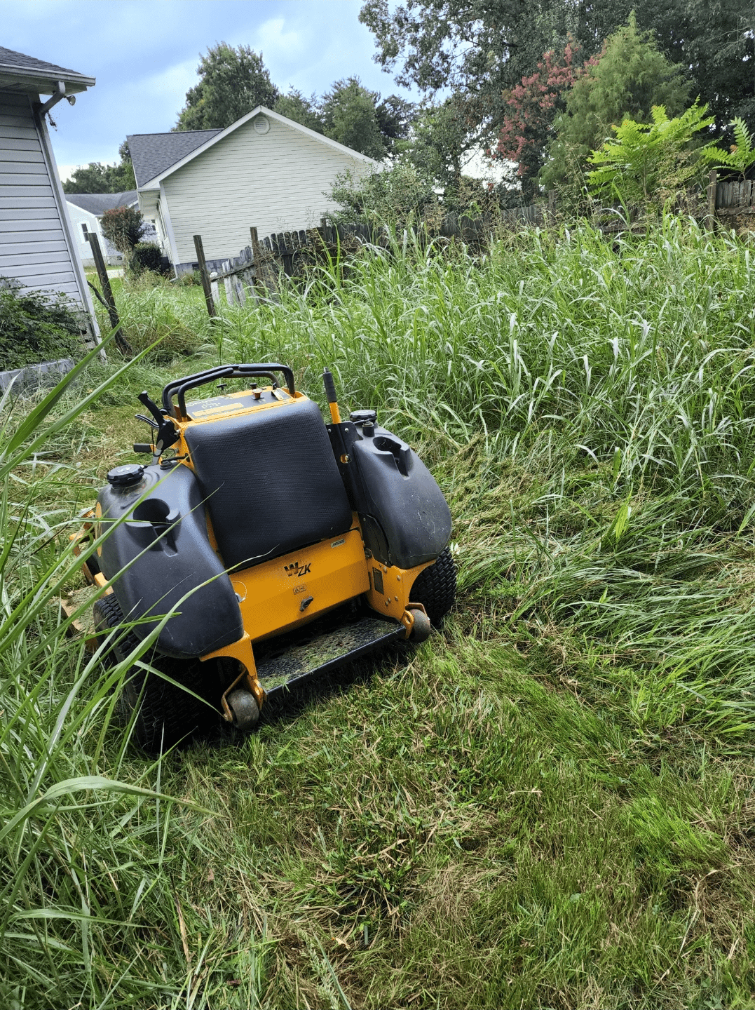 A black and orange riding lawn mower sitting in a severely overgrown lawn on a sunny day