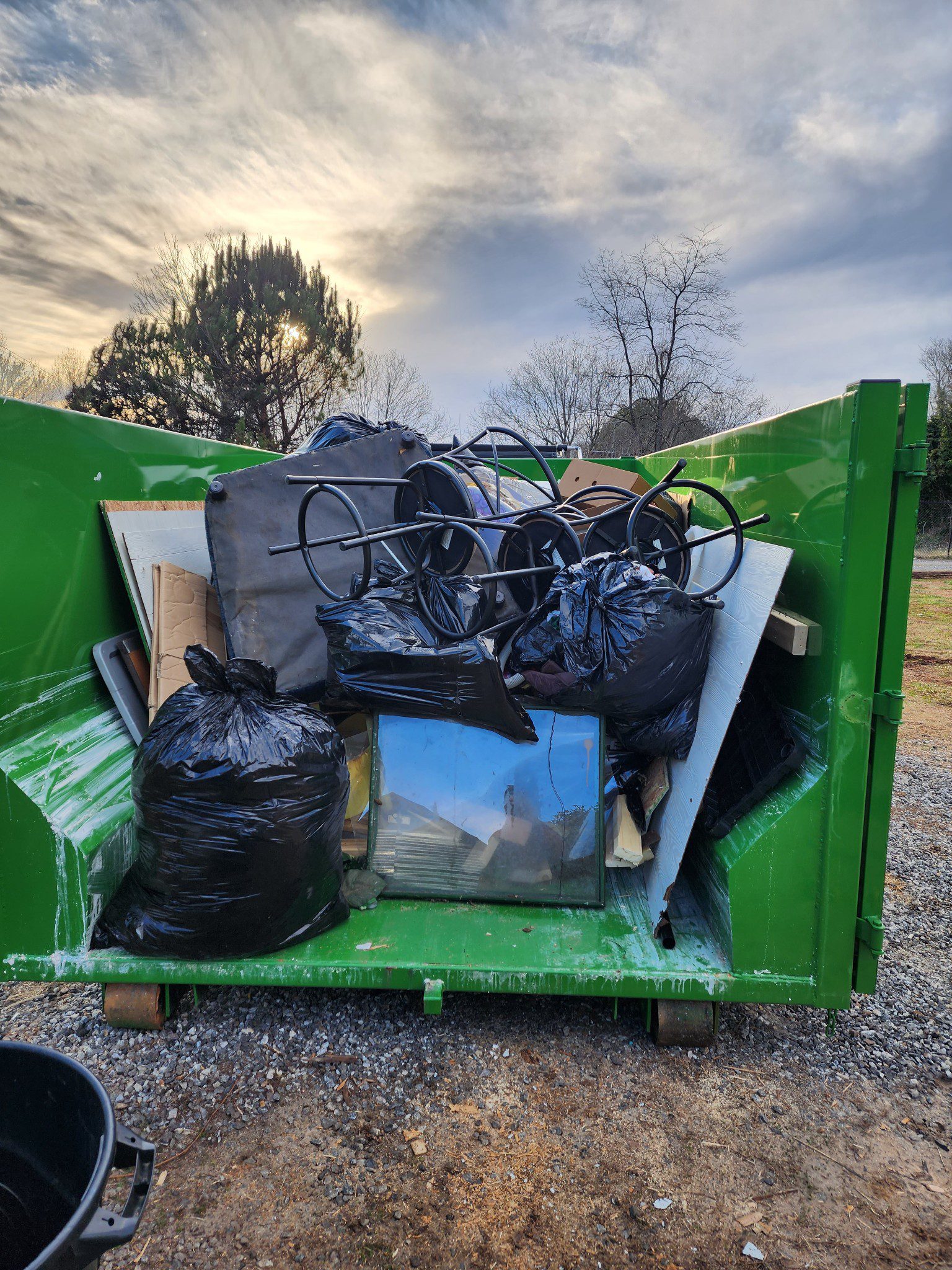A green dumpster full of household garbage in an open field at sunset